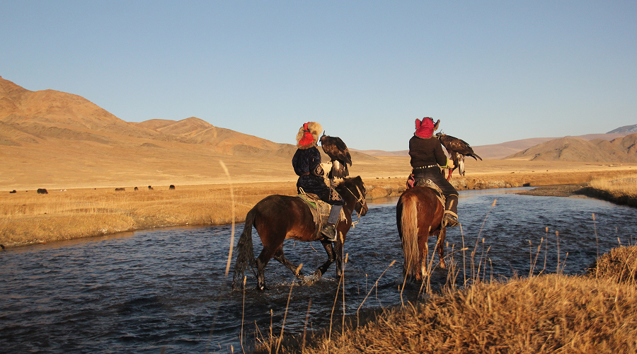 Eagle hunters riding horses across river in Altai Mountains.