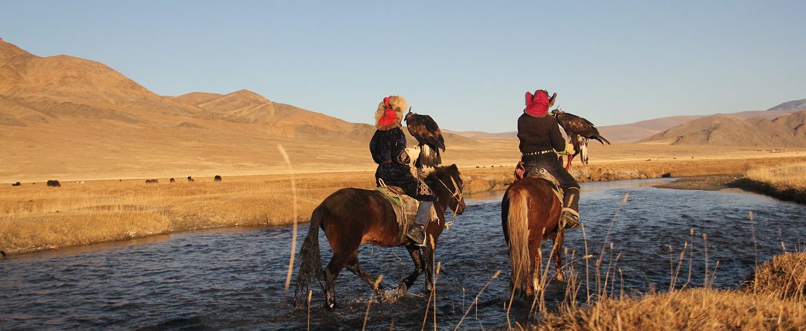 Local horsemen riding across a river with hunting eagles.