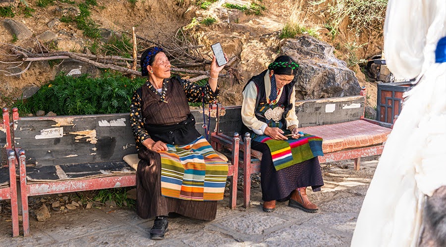 Tibetan women in traditional clothing using smartphones on a bench.
